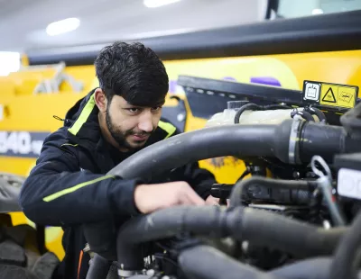 A junior technician carrying out maintenance on a yellow aerial work platform in a workshop.